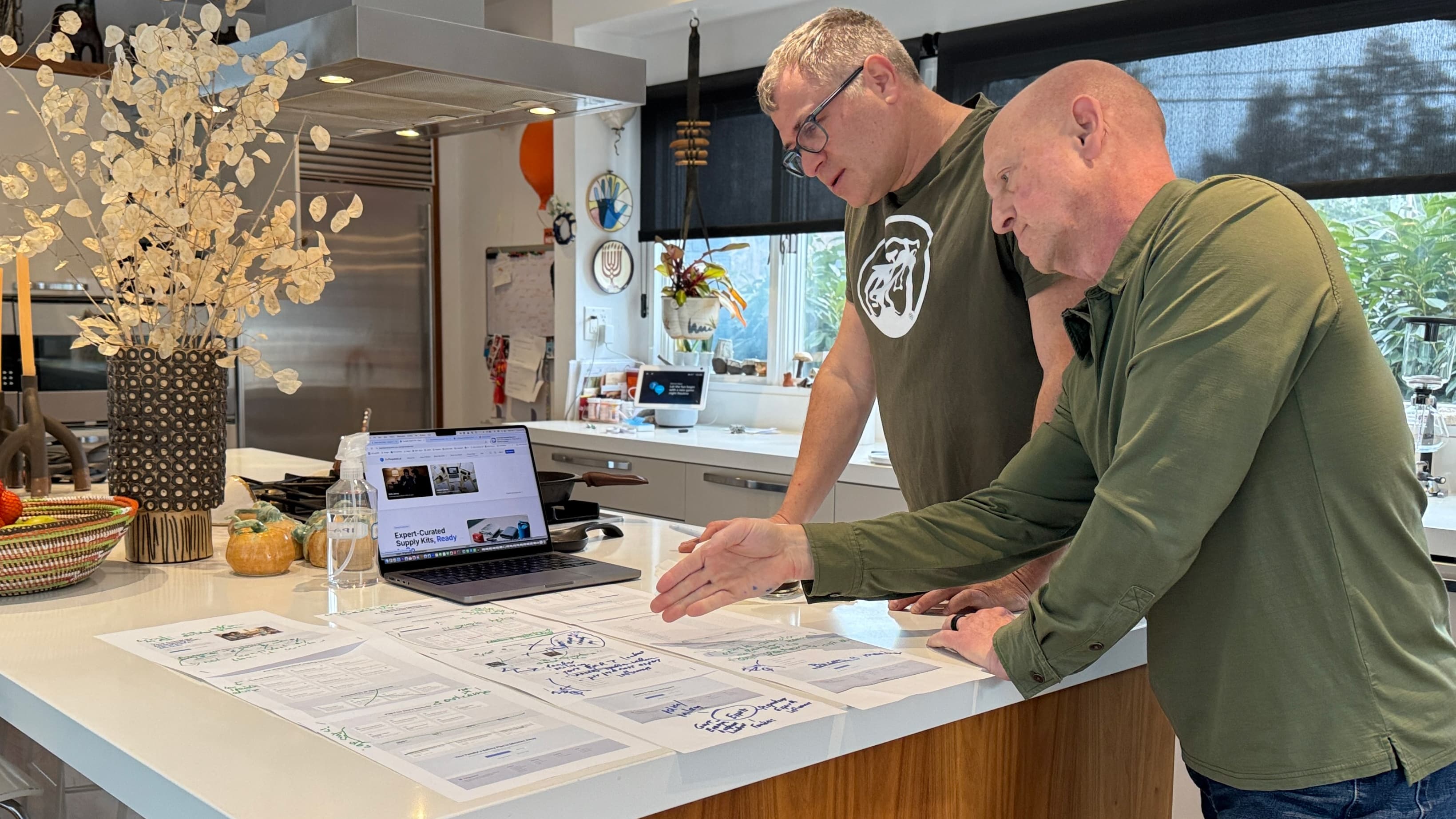 Brian and Tiran reviewing the beprepared.ai website at a kitchen island, with printed wireframes and hand-written annotations spread across the counter